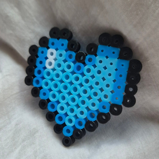 A heart-shaped bead craft made of blue and light blue Hama beads with black borders, placed on a white fabric background.