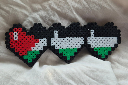 Three heart-shaped bead crafts featuring the Palestinian flag design, displayed on a white fabric background.